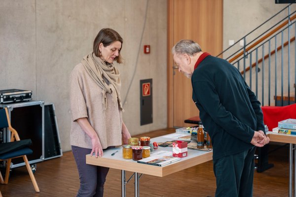 Frau Roos zeigt einem Mann ihre selbstgemachte Marmelade, die auf einem Tisch steht