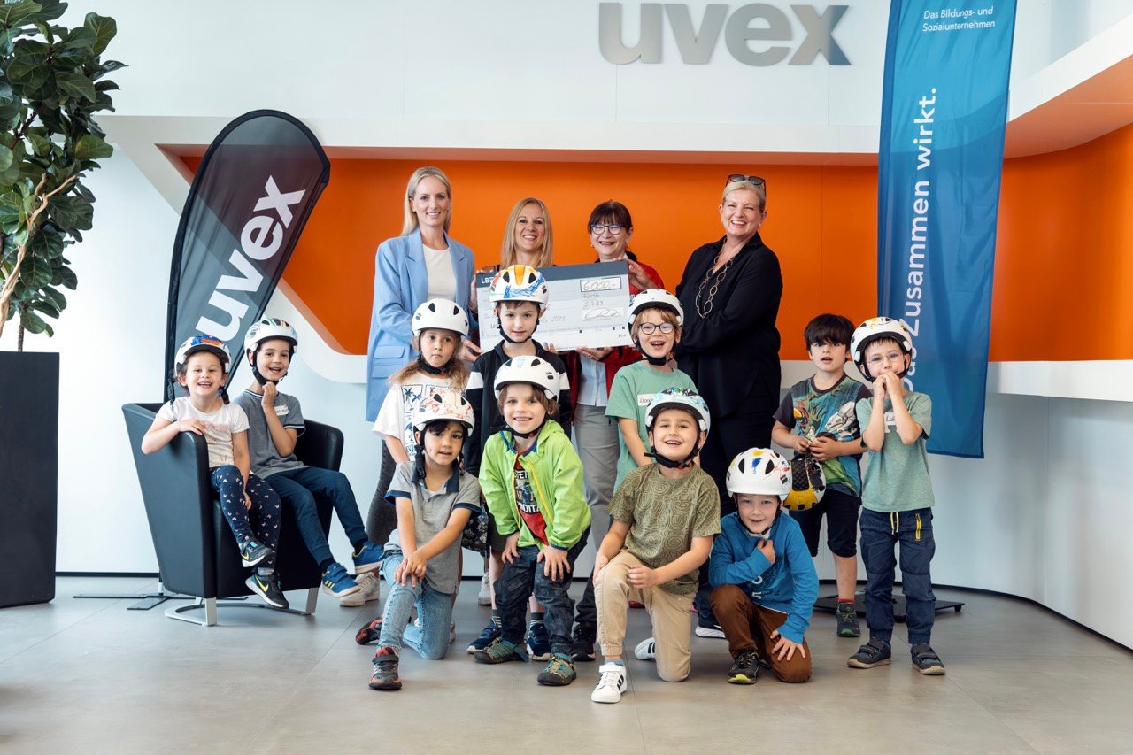 Gruppenfoto von Kindern mit Fahrradhelmen und mehreren Erwachsenen bei einer Spendenübergabe im Eingangsbereich eines Unternehmens mit uvex Branding.