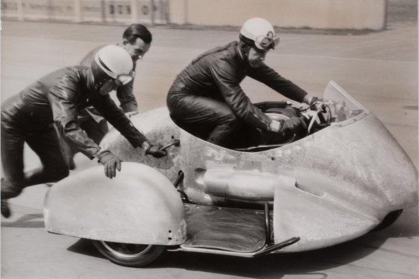 Drei Personen die ein altes Dreirad Motorrad schieben