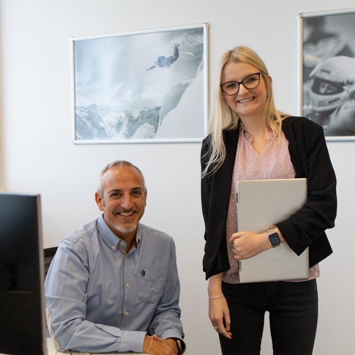 Zwei Mitarbeiter stehen im Büro und lächeln in die Kamera