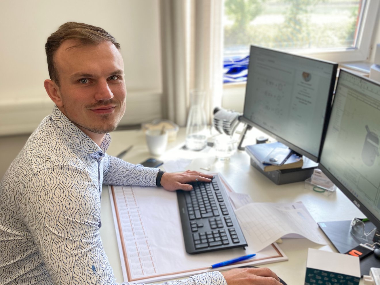 Ein Man der an zwei Bildschirmen arbeitet und in die Kamera schaut