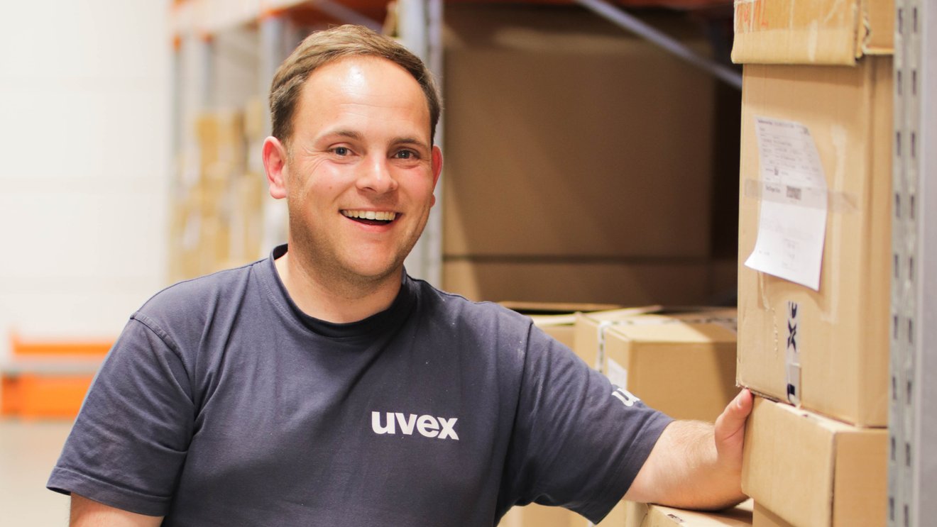 Ein junger Mann steht neben einem Regal mit uvex Kartons und lacht