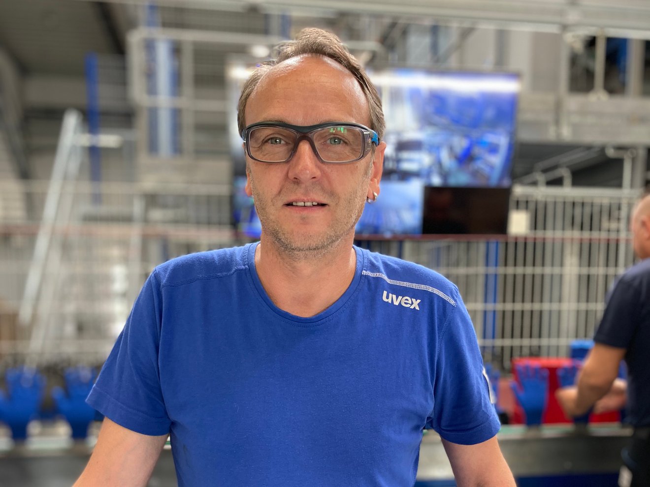 Ein Man mit blauem uvex Shirt und Schutzbrille steht in einer Fabrik